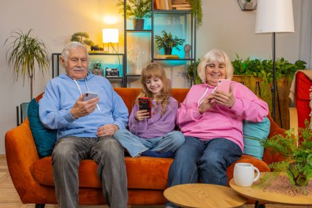 Photo for Addicted grandfather, grandmother and granddaughter using smartphones in living room at home. Smiling girl and senior couple spending leisure time sitting on sofa. Playing online games, social media. - Royalty Free Image