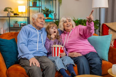 Photo for Happy Caucasian grandfather, grandmother and granddaughter eating popcorn and watching movie on sofa at home. Smiling small girl with grandparents enjoying film during weekend in living room apartment - Royalty Free Image