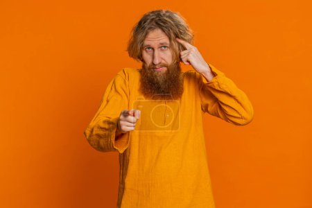 You are crazy, out of mind. Confused Caucasian man pointing at camera and showing stupid gesture, blaming asking some idiot for insane plan, bullying. Redhead young guy isolated on orange background