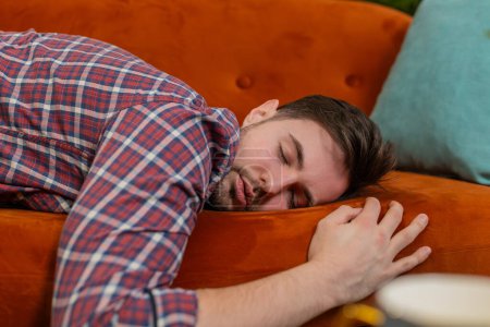 Exhausted young man in plaid shirt flopped down on sofa falls asleep. Concept of after party, tired overworked person hard day, lack of energy, breakdown. Caucasian drunk bored guy sleeping on couch.