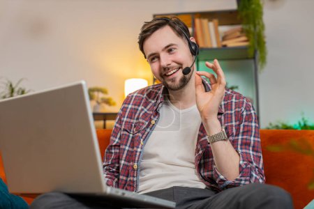 Photo for Happy excited business man helpline in plaid shirt talking to web camera headset making an online video conference call in distance. Caucasian freelancer guy uses laptop on sofa in living room at home - Royalty Free Image