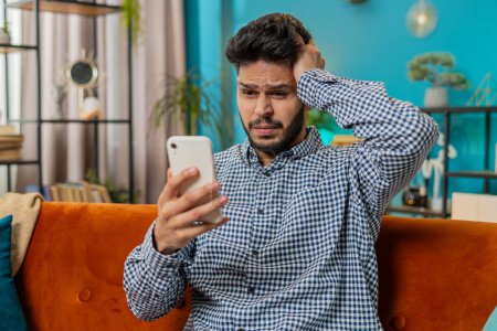 Sad displeased Indian man use smartphone typing browsing, loses becoming surprised sudden lottery game results, bad news, fortune loss fail deadline, virus. Hispanic guy at home room sitting on sofa