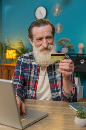 Photo for Happy senior man using bank credit card and laptop for online shopping payments at home office. Business transaction. Smiling grandpa freelancer purchases with netbook E-banking app service. Vertical - Royalty Free Image