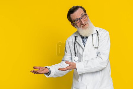 Senior elderly doctor cardiologist man raising hands asking what why reason of failure demonstrating disbelief irritation by troubles. Apothecary pharmacy old grandfather isolated on yellow background