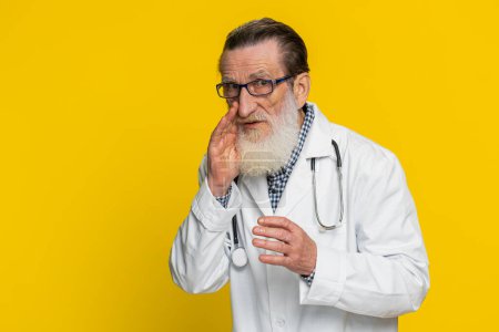Keep my secrets, silence. Senior doctor cardiologist man whisper news rumors holding hands near mouth, share gossip quiet. Grandfather telling interesting confidential information on yellow background