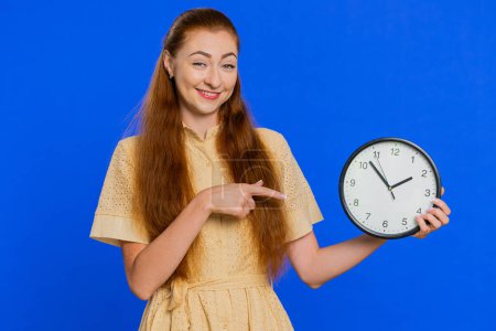 It is your time. Young adult woman showing time on wall office clock, ok, thumb up, approve, deadline, challenge, time out, success, hurry up. Redhead girl isolated on blue studio background indoors
