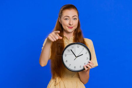 It is your time. Young adult woman showing time on wall office clock, ok, thumb up, approve, pointing finger at camera, deadline, time out, hurry up. Redhead girl isolated on blue studio background