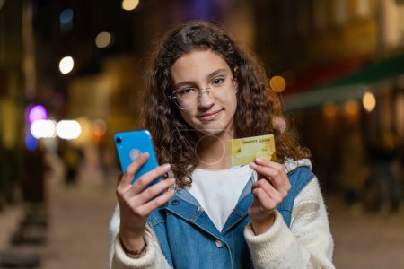 Photo for Young teenage girl using credit bank card smartphone while transferring money, purchases online shopping, order food delivery, booking hotel room. Happy woman in urban city street at night evening - Royalty Free Image