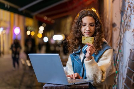Photo for Girl using credit bank card laptop while transferring money, drinking coffee purchases online shopping, booking hotel room outdoors at night. Young woman tourist sitting at table in city cafeteria. - Royalty Free Image