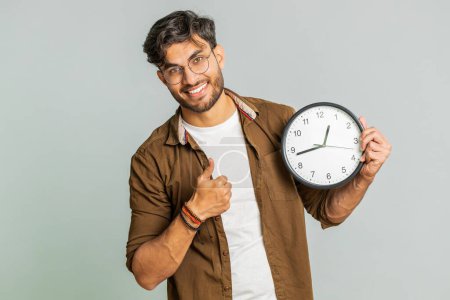 It is your time, hurry up. Indian young man showing time on wall office clock, ok, thumb up, approve, pointing finger at camera, advertisement, deadline. Arabian guy isolated on studio gray background