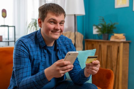 Hombre sonriente usando tarjeta bancaria de crédito y teléfono inteligente durante la transferencia de dinero, compras en línea sin efectivo pedido entrega de alimentos en el apartamento de casa en el interior. Feliz joven se sienta en la habitación en el sofá.
