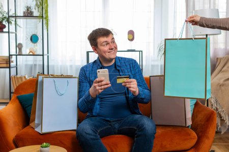Photo for Happy shopaholic consumer man sitting with shopping bags at apartment making online order, payment with credit plastic bank card. Young guy receives order on couch at home. Discount sale on internet. - Royalty Free Image