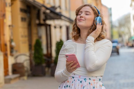 Photo for Happy relaxed overjoyed redhead young woman in wireless headphones listening energetic disco music in smartphone dancing outdoors. Girl in long dress walking in urban city street. Town lifestyles. - Royalty Free Image