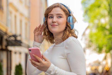 Photo for Happy relaxed overjoyed redhead young woman in wireless headphones listening energetic disco music in smartphone app dancing outdoors. Red hair girl walking in urban city street. Town lifestyles. - Royalty Free Image