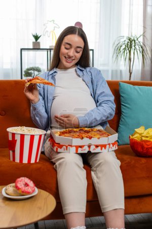 Pregnant woman eating unhealthy junk food pizza, popcorn crisp sitting on sofa watching TV movie in home cozy room. Fast fatty food during pregnancy in apartment. Hunger, change in taste preferences