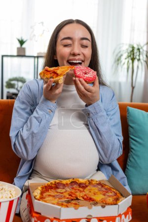 Pregnant happy smiling woman eating unhealthy junk food pizza, popcorn crisp sitting on sofa in home living room. Fast fatty food during pregnancy in apartment. Hunger, change in taste preferences