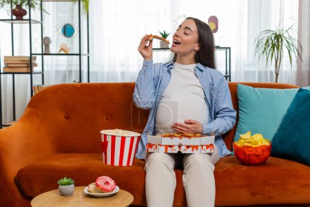 Pregnant woman girl eating unhealthy junk food pizza, popcorn crisp sitting on couch watching TV movie in home room. Fast fatty food during pregnancy in apartment. Hunger, change in taste preferences