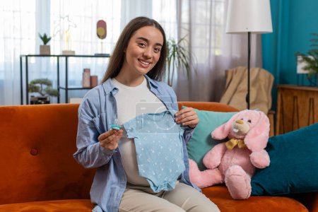 Pregnant woman looking at baby cloth and pacifier. Future mother enjoys maternity, look at baby cloth with tenderness sitting on couch at home. Preparing pregnancy, expecting baby, childbirth concept