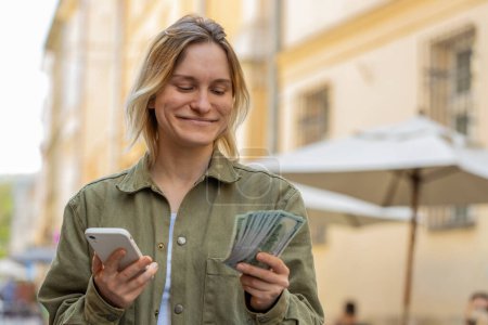 Glückliche junge Frau, die per Smartphone-Taschenrechner-App Geld in bar zählt, zufrieden mit dem Einkommen für geplante Weihnachtsgeschenke. Kaukasisches Mädchen auf der sonnigen Straße der Großstadt. Stadtleben im Freien.