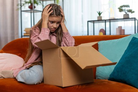 Angry dissatisfied shopper child girl unpacking parcel feeling upset and confused with wrong mistake delivery from an online store, bad quality broken purchase at home. Kid showing thumb down on sofa.