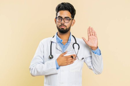 I swear to be honest. Sincere responsible Indian young doctor cardiologist man raising hand to take oath, promising to tell truth, keeping hand on chest. Apothecary pharmacy guy on beige background
