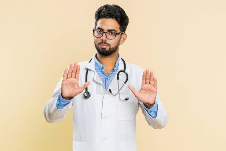 Hey you, be careful, take care. Strict Indian young doctor cardiologist man warning with admonishing hands gesture, giving advice to avoid danger. Arabian apothecary pharmacy guy on beige background