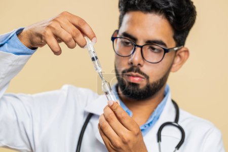 Indian young doctor cardiologist man holds syringe needle and ampoule tube with medical vaccine medicine treatment injection ready to use. Immunization. Arab scientist guy isolated on beige background