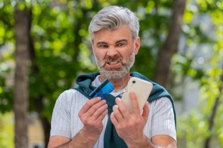 Shocked Caucasian man trying to pay online shopping with smartphone blocked credit card. Annoyed bearded mature tourist on street buying bank refuse problem unsuccessful payment lack of money balance.