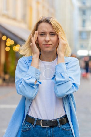 Displeased exhausted Caucasian woman rubbing temples to cure headache problem suffering from tension migraine stress. Young lady tourist grimacing pain high blood pressure stands on city urban street