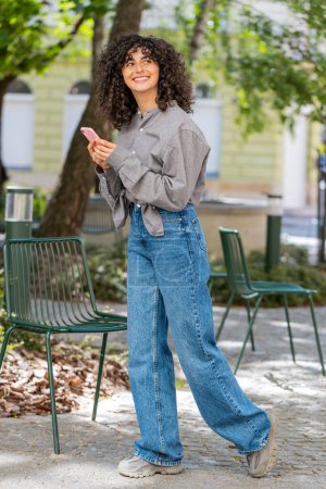 Young tourist woman uses smartphone typing text messages looking for search a way on map in mobile navigator app outdoors. Caucasian curly hair girl walking passes by urban city street park. Vertical