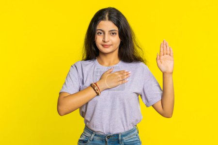 I swear to be honest. Sincere responsible Indian woman raising hand to take oath promising to be honest, to tell truth, keeping hand on chest justice. Arabian girl isolated on yellow studio background