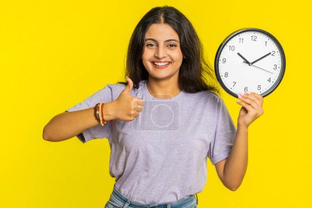 It is your time, hurry up. Indian woman showing time on wall office clock, ok, thumb up, approve, pointing finger at camera, advertisement, deadline. Arabian girl isolated on studio yellow background