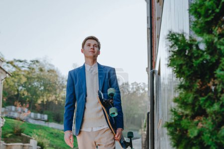 Foto de Sonriente joven hombre de negocios guapo con traje mensajería celebración longboard caminar calle - Imagen libre de derechos