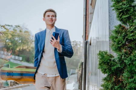Foto de Sonriente joven hombre de negocios guapo usando mensajería de traje usando el teléfono celular sosteniendo longboard - Imagen libre de derechos