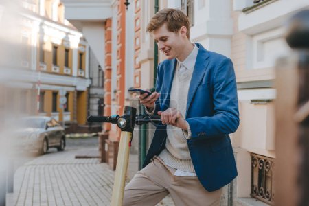 Téléchargez les photos : Jeune homme en costume utilisant smartphone pour scooter électrique louer dans la rue de la ville. Concentration sélective. - en image libre de droit