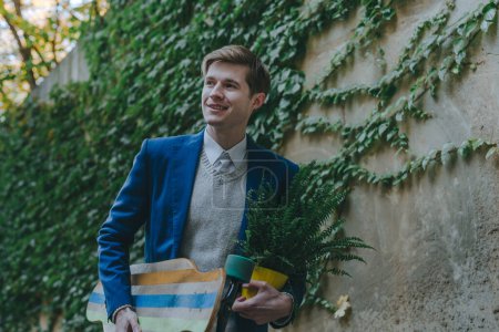 Photo for Smiling handsome young man businessman wearing suit holding fern plant and longboard near wall covered with ivy outdoor. - Royalty Free Image