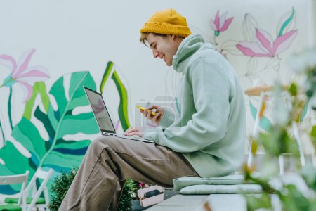 Téléchargez les photos : Jeune homme indépendant travaillant sur un ordinateur portable à l'aide d'un smartphone assis dans les escaliers de la rue. Concentration sélective. - en image libre de droit