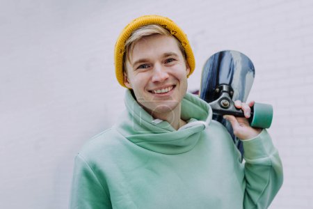 Foto de Sonriente joven guapo sosteniendo longboard cerca de pared de ladrillo al aire libre. - Imagen libre de derechos