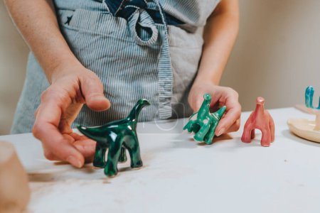 Photo for Young woman ceramist holding small colorful ceramic dinosaur figurines in the workshop. Close up, selective focus. - Royalty Free Image