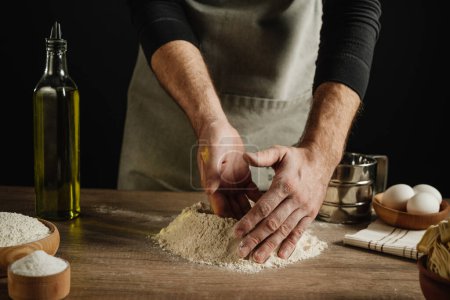 Photo for Unrecognizable man kneads the dough on a wooden background against a dark wall. Close-up of male hands forming flour. Step 1 of making homemade noodles. Selective focus. - Royalty Free Image