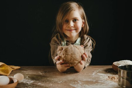 Téléchargez les photos : Petite fille dans un tablier tenant une pâte contre un mur sombre. L'enfant prépare des gâteaux faits maison ou des nouilles. Concentration sélective. - en image libre de droit