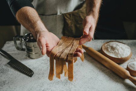 Photo for Unrecognizable man in an apron holding dough cut into strips on a white table against a dark wall. Close-up of male hands preparing homemade noodles. Selective focus. - Royalty Free Image