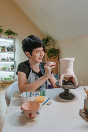 Photo for Female craft person potter painting a vase sitting at her workshop. Selective focus. - Royalty Free Image