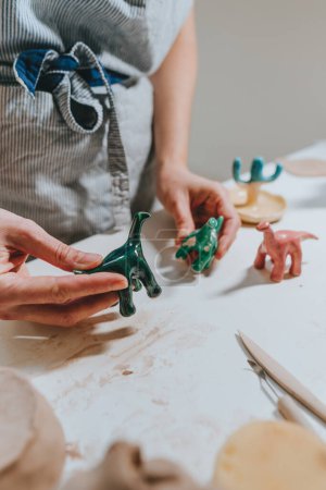 Foto de Mujer joven ceramista sosteniendo pequeñas figuras de dinosaurios de cerámica de colores en el taller. Primer plano, enfoque selectivo. - Imagen libre de derechos