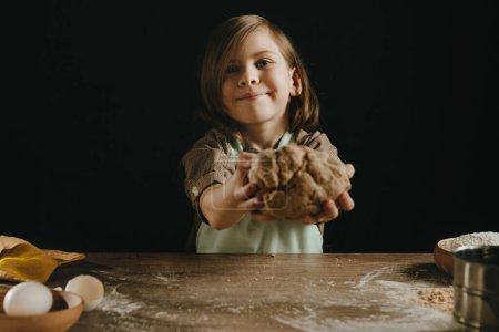 Téléchargez les photos : Petite fille dans un tablier tenant une pâte contre un mur sombre. L'enfant prépare des gâteaux faits maison ou des nouilles. Concentration sélective. - en image libre de droit