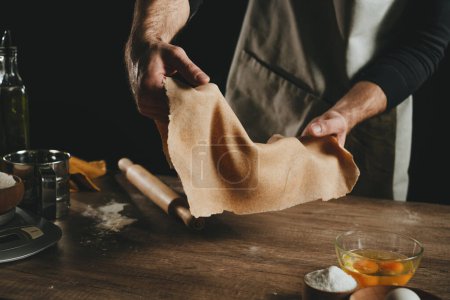 Photo for Unrecognizable man holding a thin dough against a dark background. Close-up of male hands preparing homemade noodles. Close-up of male hands preparing homemade noodles. Selective focus. - Royalty Free Image