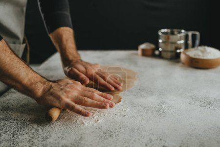 Photo for Unrecognizable man rolling out the dough with a rolling pin on black background. Close-up of male hands prepairing homemade noodles. Selective focus. - Royalty Free Image