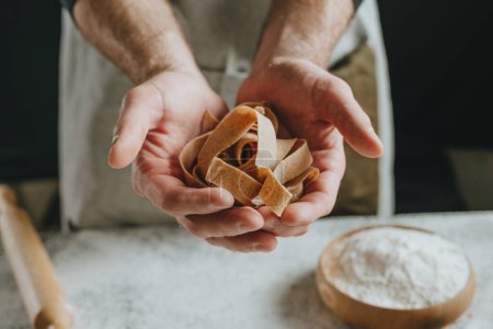 Photo for Unrecognizable man in an apron holding dough cut into strips on a white table against a dark wall. Close-up of male hands preparing homemade noodles. Selective focus. - Royalty Free Image