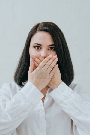 Photo for Young brunette woman in a white shirt closing her mouth with her hands against white background. Selective focus. - Royalty Free Image
