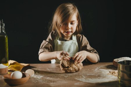 Photo for Little girl wearing apron kneading dough on wooden background against dark wall. Child prepairing homemade bakery or noodles. Selective focus. - Royalty Free Image